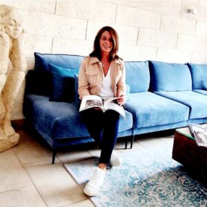 Claudia Müller | Sitzend auf einer Couch lächelnd mit einem Home Staging Prospekt in der Hand.